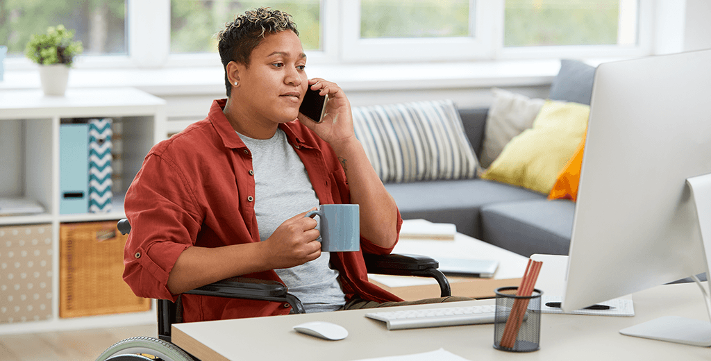 Image of a woman of colour wearing a terracotta colour shirt and holding a mug of coffee. She is a wheelchair user and is on the phone at her home computer.