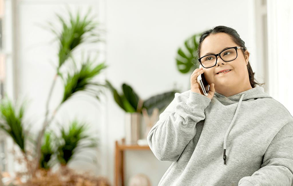 Image of a woman with Down Syndrome talking on her mobile, wearing a grey jumper and glasses, image has green house plants in the background.