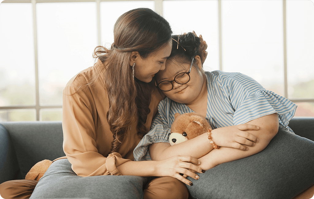 Image of a young woman with intellectual impairment holding a teddy bear and resting her head on her mother's shoulder.