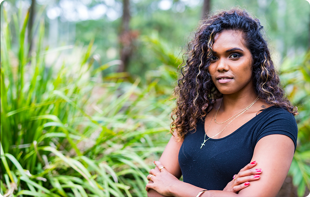 Young Indigenous woman with long curly hair wearing a black t-shirt, standing in a garden with her arms crossed.