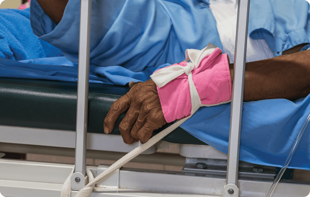 Photo of an older indigenous person in a blue hospital gown lying in a bed. Photo is close up on their hand which is in a bright pink fabric restraint tied to the bed rail.