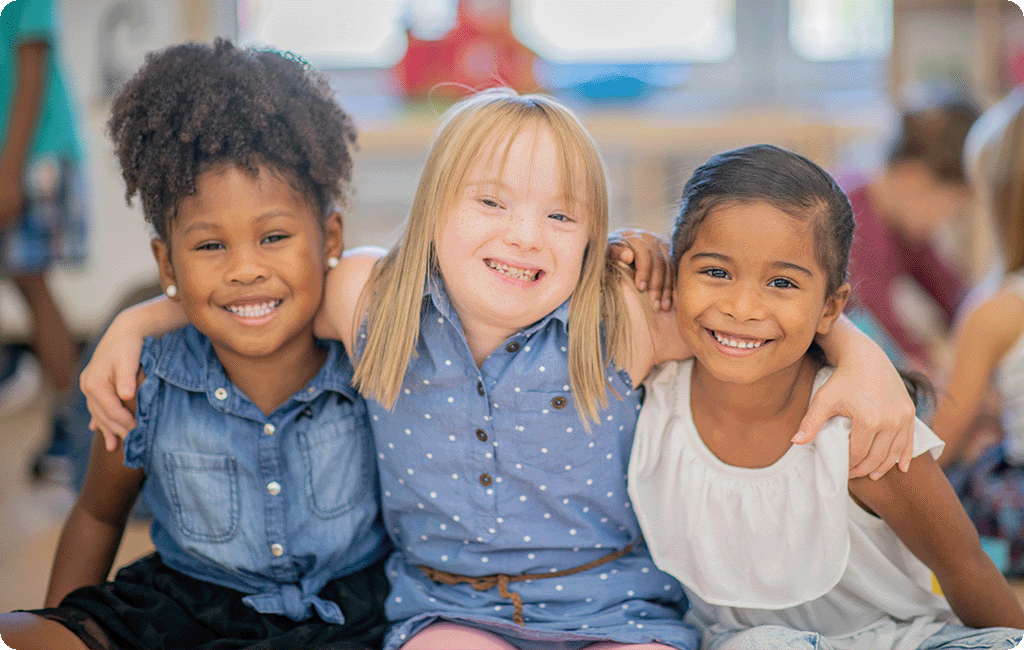 3 young girls in school, 2 girls of colour on either side of a Caucasian girls with disability, they have their arms around each other and are all smiling.