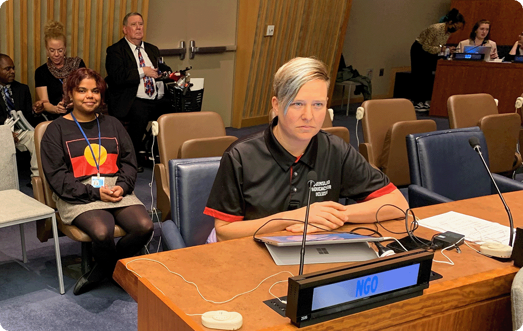 Matilda Alexander sitting at bench in front of microphone and sign that says "NGO" at the United Nations Conference of States Parties General Debate. Naraja Clay and others are sitting behind Matilda.