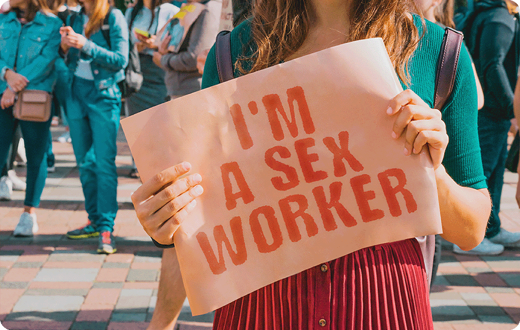 Woman holding a sign that says "I'm a sex worker" with a group of people in the background.