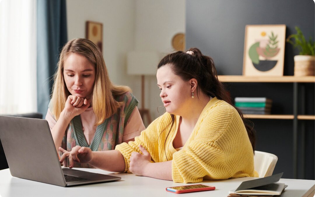 Woman with Down Syndrome wearing a yellow jumper, she is working on a laptop at a table with a blonde woman wearing a pink top.