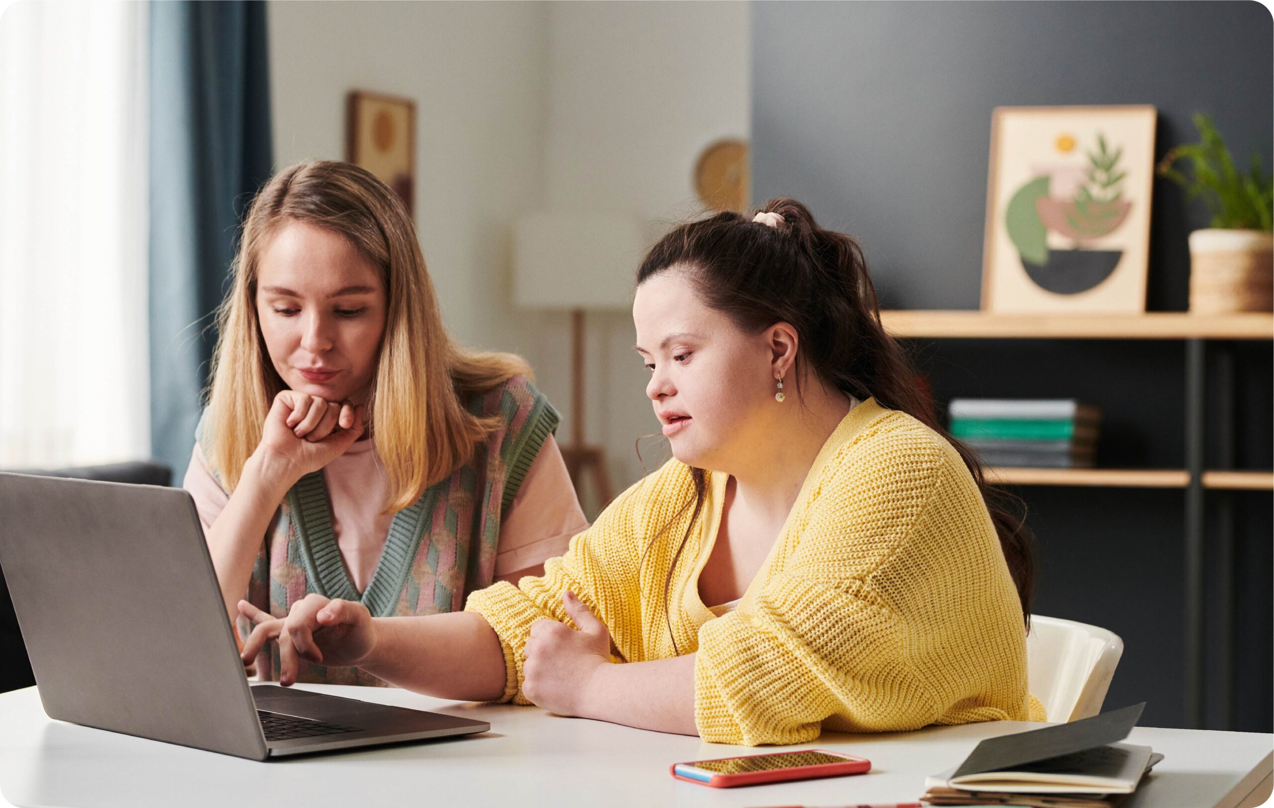 Woman with Down Syndrome wearing a yellow jumper, she is working on a laptop at a table with a blonde woman wearing a pink top.