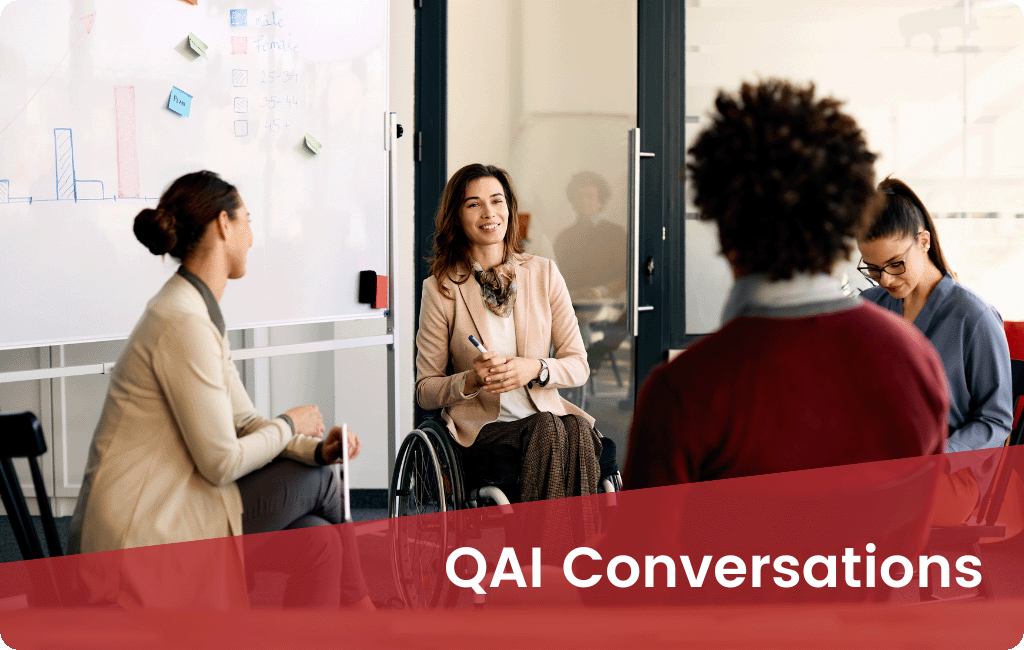 Four people, one is a wheelchair user, in a bright office sitting in a circle talking. Red overlay with white text says "QAI Conversations".