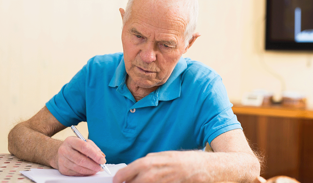 Elderly man in bright blue t-shirt filling in a form at a table.
