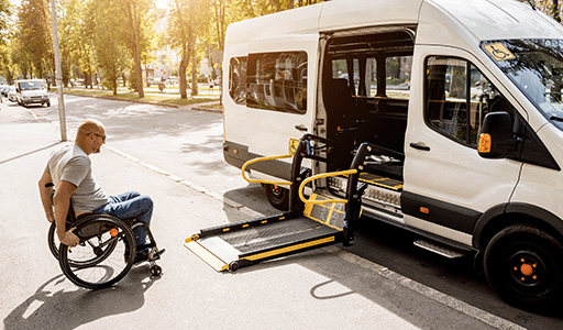 Man using wheelchair about to move onto a chair lift to get into a white van on a sunny street lined with trees.