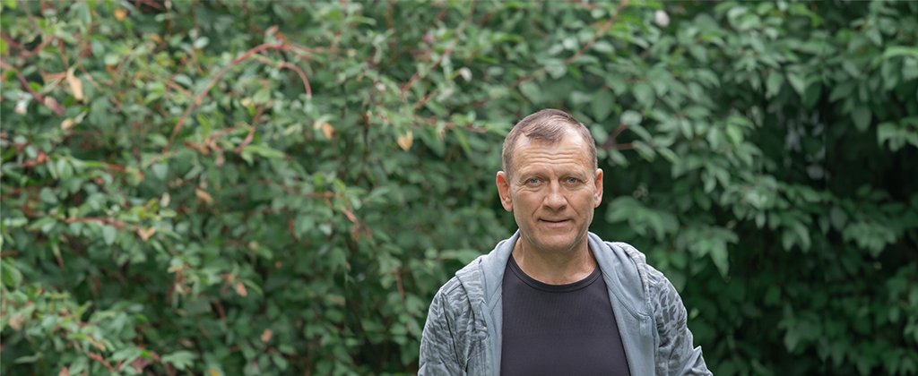 Middle-aged Caucasian man looking at camera, he has balding hair and is wearing a dark T-shirt and hoodie. Background is full of out green leaves.