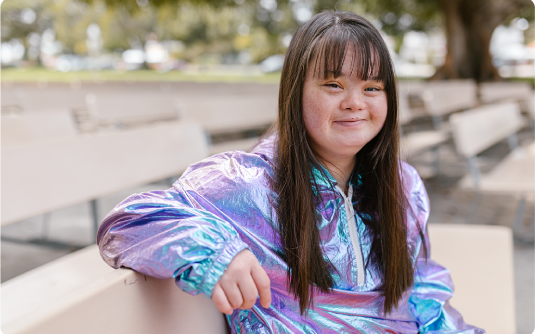 Young woman with Down Syndrome wearing an shiny purple and blue jacket, she is sitting on an outdoor auditorium bench and has a slight smile on her face.