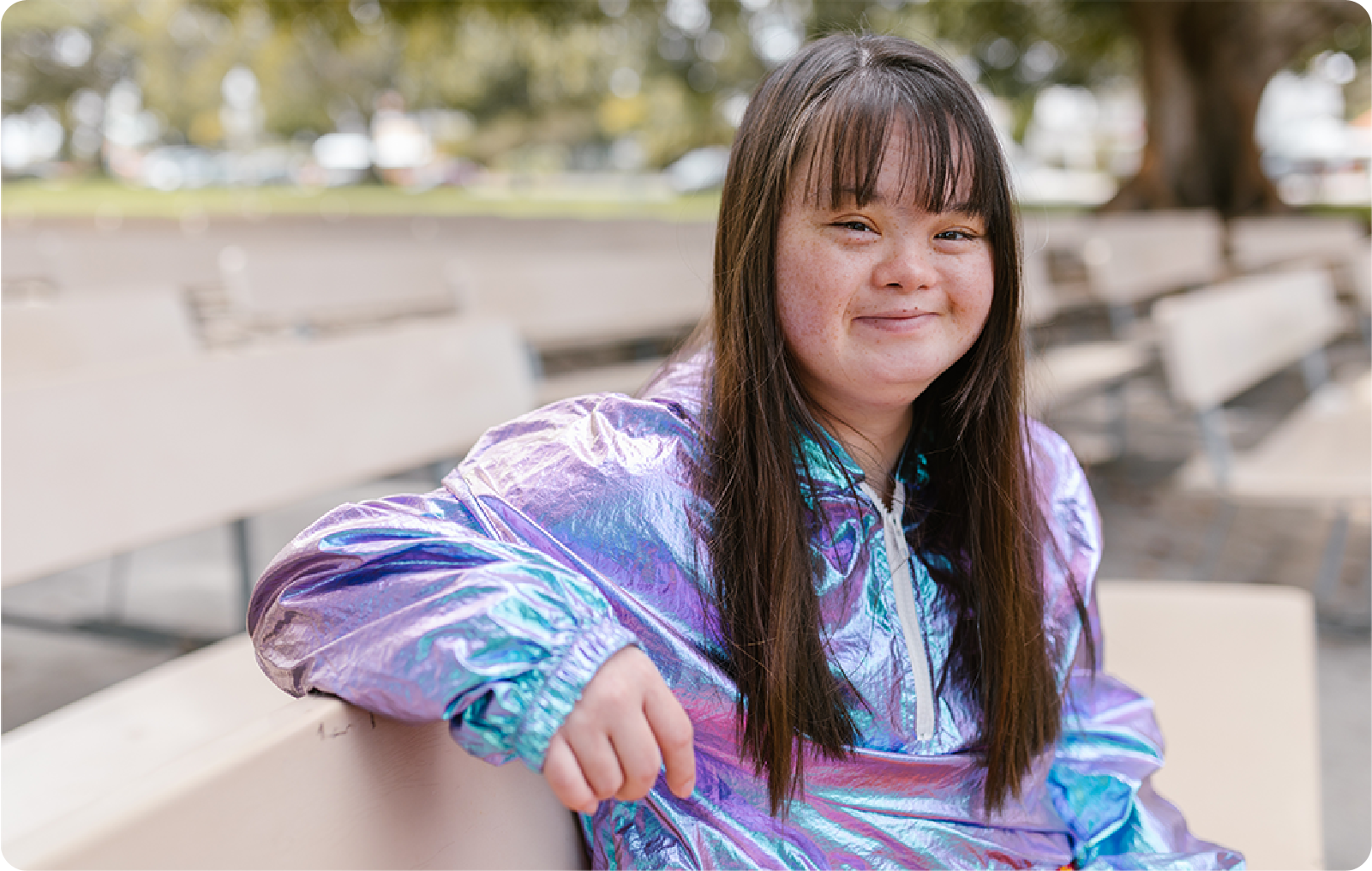 Young woman with Down Syndrome wearing an shiny purple and blue jacket, she is sitting on an outdoor auditorium bench and has a slight smile on her face.