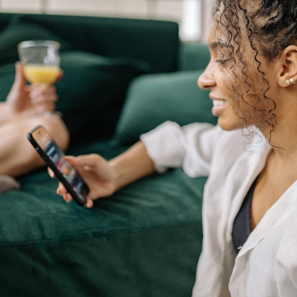Woman of colour sitting on floor against green sofa scrolling on her phone, friend on sofa in background.