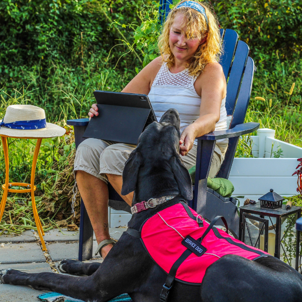 Woman sitting on patio with her tablet on her lap and service dog at her feet.