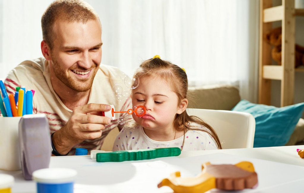An adult helps a child blow bubbles while playing at a desk.